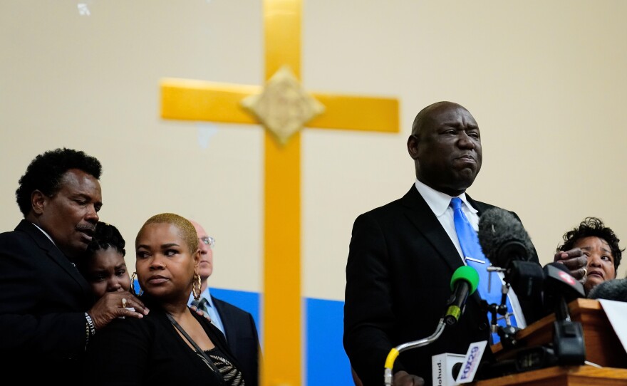 Ben Crump stands with several other people at a podium in front of a yellow cross on the wall. 