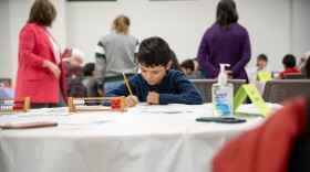 young boy sits at table with simple abacus in front of him, holding pencil and looking down at paper.