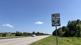 A wide shot of Highway 54 in rural Mid-Missouri. A "West 54" road sign stand in the ditch. The grass on the side of the road is green. A white sedan travels along the highway in the distance. 