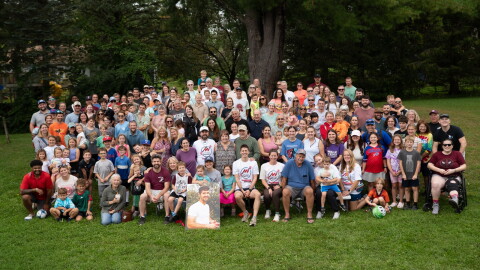 This is a group of people posing in a park in front of a tree. They have a poster with a picture of Nick Henry, who passed away after a battle with cancer. 