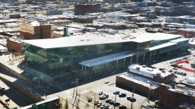 A large building with a green, glass exterior sits in the middle of a city block, surrounded by other smaller buildings.