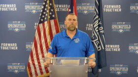 A man wearing a blue shirt stands at a glass podium with flags behind him.