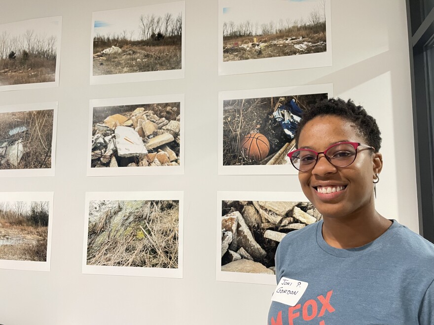 A woman in glasses smiles at the camera. She is standing in front of photographs of dilapidated buildings and nature reserves.