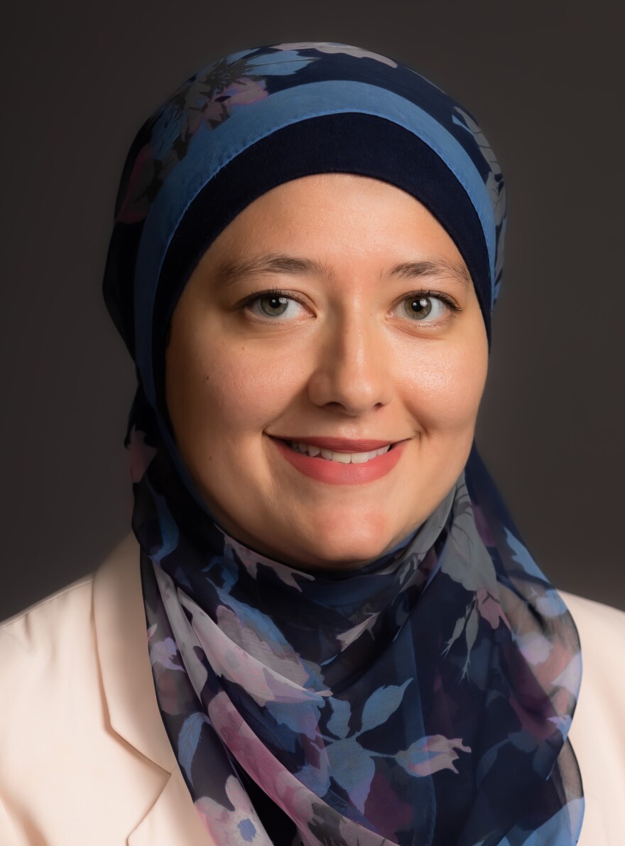 A woman wearing a blue patterned hijab and a light-colored blazer, smiling with a dark backdrop.