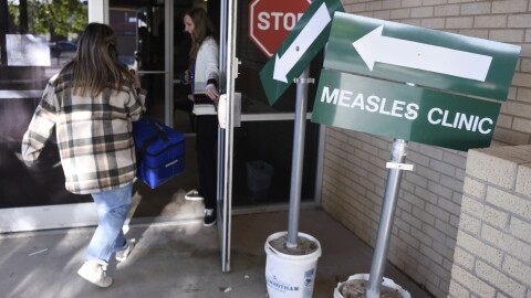 A woman holds a door open as another woman with a flannel shirt walks in. Next to the door is a sign that reads "Measles Clinic"