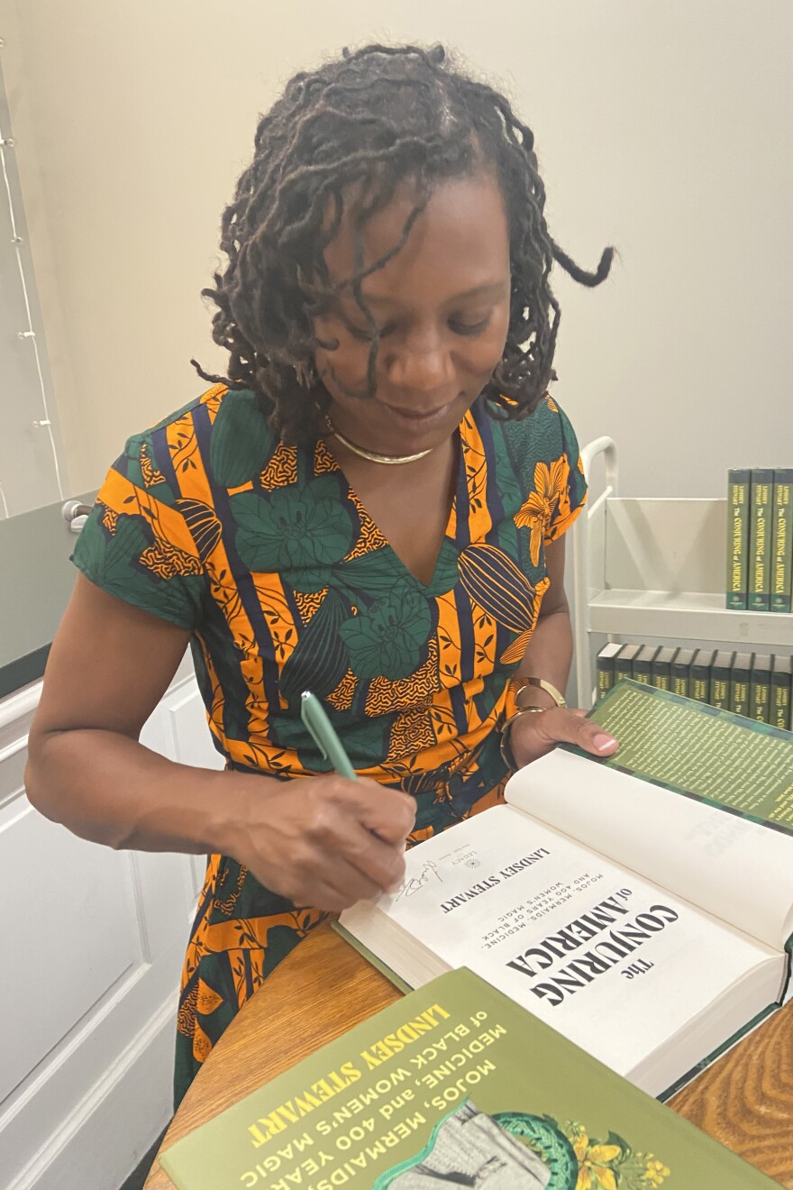 Lindsey Stewart wears a green, black and orange patterned dress, a gold necklace and gold bangles to a book signing event. On the table in front of her is a stack of her book, "The Conjuring of America: Mojos, Mermaids, Medicine, and 400 Years of Black Women's Magic," with one open in front of her to the title page, where Stewart is signing her name.