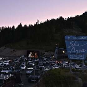 Dozens of vehicles are parked at a drive-in movie screening at Mount Ashland Ski Area at dusk, with the ski area sign in the foreground and forested mountain slopes in the background.
