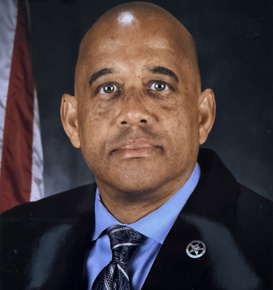 Close up of man in a dark blue suit and tie seated in front of a U.S. flag