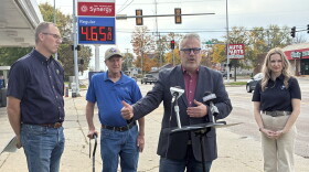 A man in a blazer speaks at a podium with two microphones, flanked by a man in a blue polo, an older man with a cane, and a woman in a black polo, with a gas station price sign in the background.