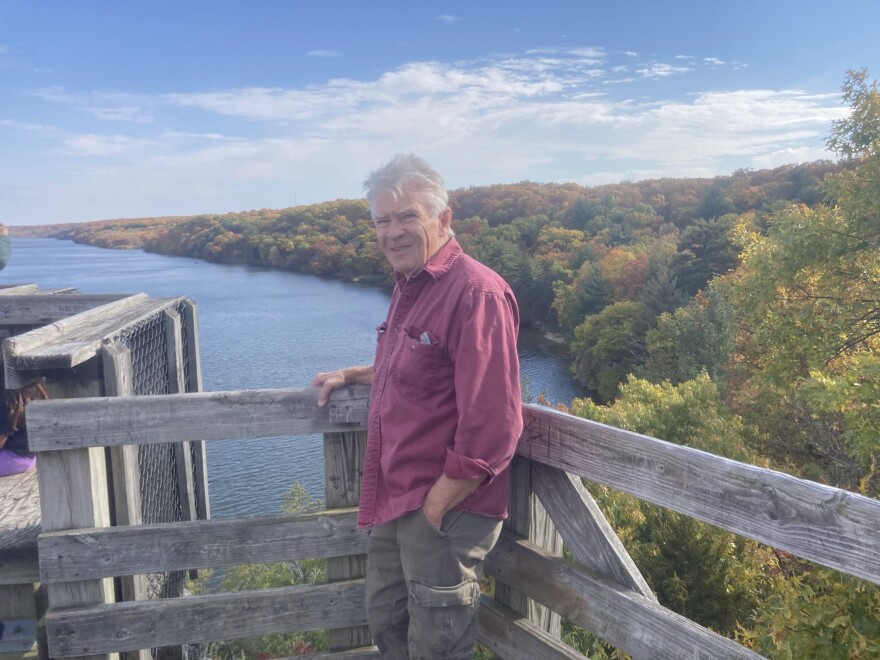 A man in a red shirt and cargo pants stands leaning against a fence. Behind him is a river and line of trees.