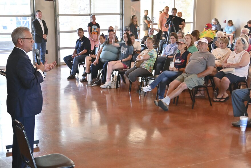 A man wearing a blue suit stands at left holding a microphone and gesturing with his left hand. To the right, people seated in chairs listen inside a large meeting space.