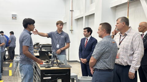 A group of professionally dressed, middle-aged men listen to two high-school-aged boys talk about what they're learning in a warehouse-style classroom.