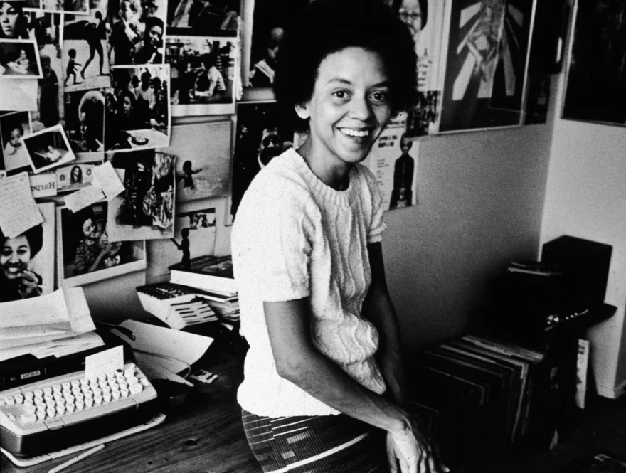 American poet Yolanda Cornelia 'Nikki' Giovanni leans on her desk beside a typewriter, in front of a wall decorated with photos, 1973.