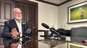 Man with gray hair and beard wearing a dark suit jacket and sitting at a table to the left with microphones in front of him