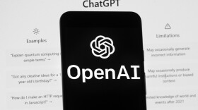 The OpenAI logo is seen on a mobile phone in front of a computer screen which displays the ChatGPT home screen.
