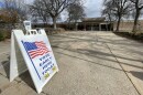 A white sandwich board sign rests on the concrete outside a building. On the sign is an illustration of an American flag, waving. The sign says VOTE EARLY HERE TODAY in blue letters, and 9a - 5p underneath