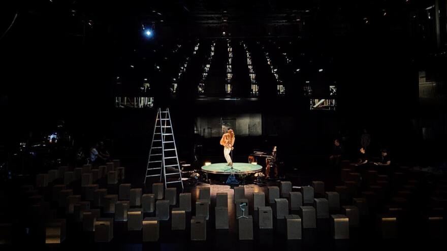 A woman performs on a small circular stage holding an acoustic guitar while surrounded by speakers and an A-frame ladder in an otherwise-empty theater.