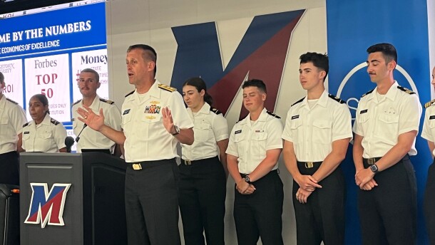 SUNY Maritime College President and retired Rear Adm. John Okon speaks in front of a podium with the letter "M" for SUNY Maritime College. Cadets in uniforms with white shirts and black pants stand in a line behind him. 