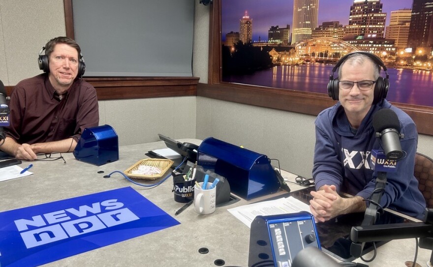 Two smiling men wearing headphones sit at a table in a radio talk studio: the man at left has short brown hair and is wearing a maroon button-down shirt; the man at right has short dark hair and is wearing glasses and a navy blue hoodie with "WXXI" in white lettering