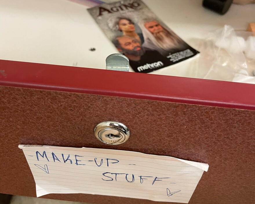 In the foreground, the front of an open drawer with an index card attached that says "make-up stuff" in handwritten letters. Inside the drawer, out of focus, is a label with pictures of people in gray hair and beards that says "Aging" at the top.