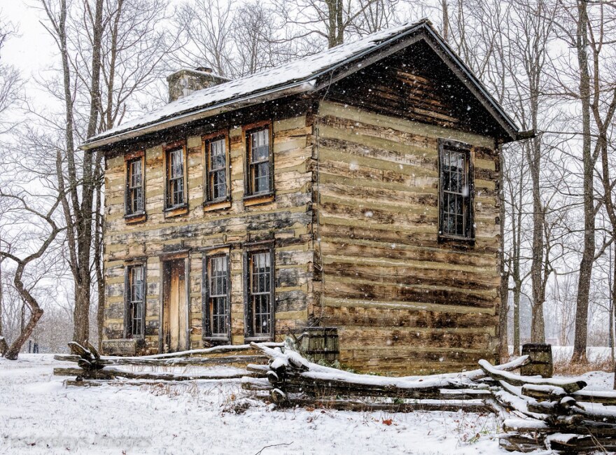 two story log house seen through snowfall with snow covered ground and bare trees