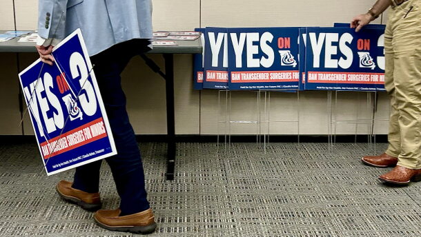 Two people from the waist down, one on either side of a conference room, are holding "Yes on 3" yard signs.