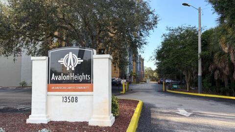 A sign that says "Avalon Heights" in front of the building's driveway. Behind it are the gates and some buildings in the distance.