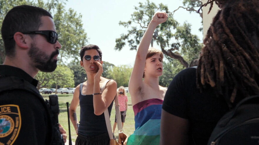 One student in a rainbow dress yells with a first raised above her head. Another in a black shirt is mid-shout.
