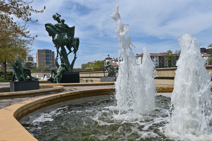 Two large spouts of water erupt from a concrete pool. A bronze statue of angels riding a horse and other bronze statues lie behind it near a cityscape.