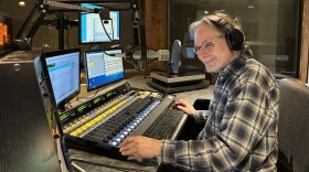 Studio One host Mark Simmet wears a flannel shirt and headphones as he sits at a soundboard with multiple computer screens in front of him.