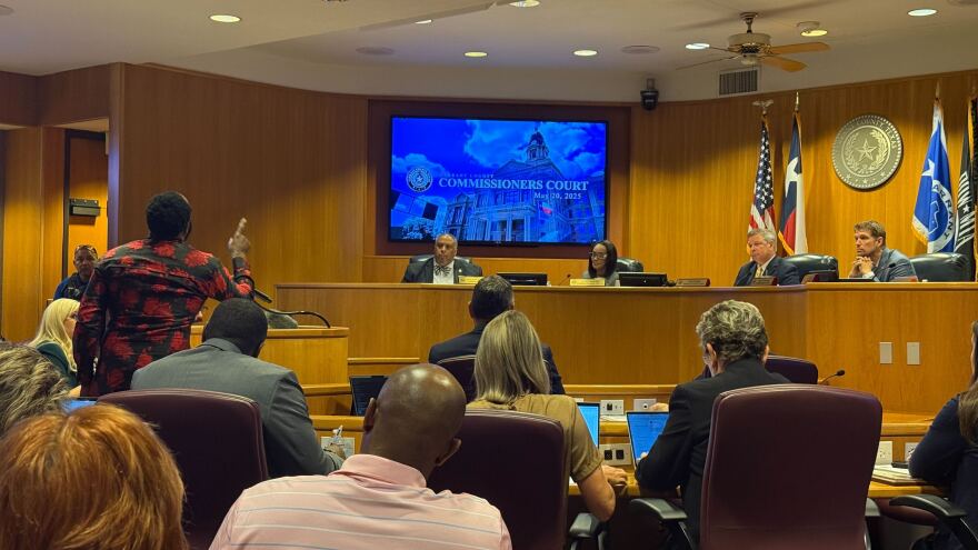 A photo of Bishop Mark Kirkland, a Black man wearing a long black and red shirt, stands at a podium in a crowded government meeting room. He is seen from behind and pointing at elected officials at the dais as he speaks.