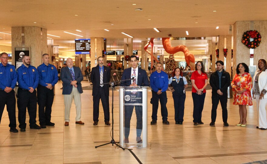 A wide-shot of a man in a suit at a podium. A group of people, some wearing uniforms, stand behind him, along with a giant fiberglass and resin flamingo.