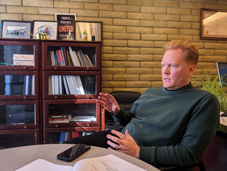 A man wearing a green sweater sits in a chair and gestures with his hands as he looks to the left. There is a brown bookshelf in the background with various binders and posters inside.