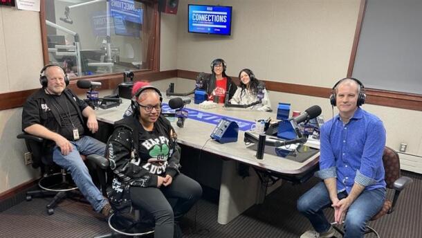 Five smiling people wearing headphones sit around a table in a radio talk studio: a man front left has short white hair and a white beard and mustache and is wearing a black t-shirt, black short-sleeved button-down shirt, jeans, and brown shoes; a young woman front center has pink hair in a ponytail and is wearing glasses, a black t-shirt that reads "HAPPY GO LUCKY" with a green, white, and orange shamrock, a black zip-up hoodie with white lettering, black pants, and black shoes; a woman back left has long blue hair and is wearing glasses, a red t-shirt that reads "FIND YOUR SPARK" in white lettering, and and black zip-up hoodies; a young woman back center has long black hair and is wearing a white sweatshirt; a man at right has short dark hair and is wearing a blue patterned button-down shirt, jeans, and sneakers