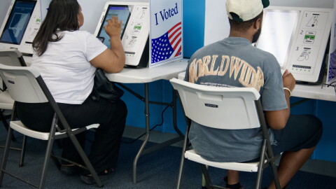 Interior photo that shows two people sitting in folding chairs with their backs to the camera. They appear to be making selections on electronic screens that are voting machines.