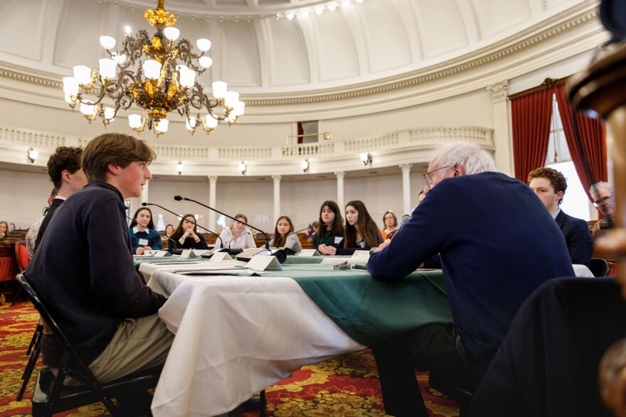 High school students sit around a table alongside US Senator Bernie Sanders, who is seated at the head of the table.