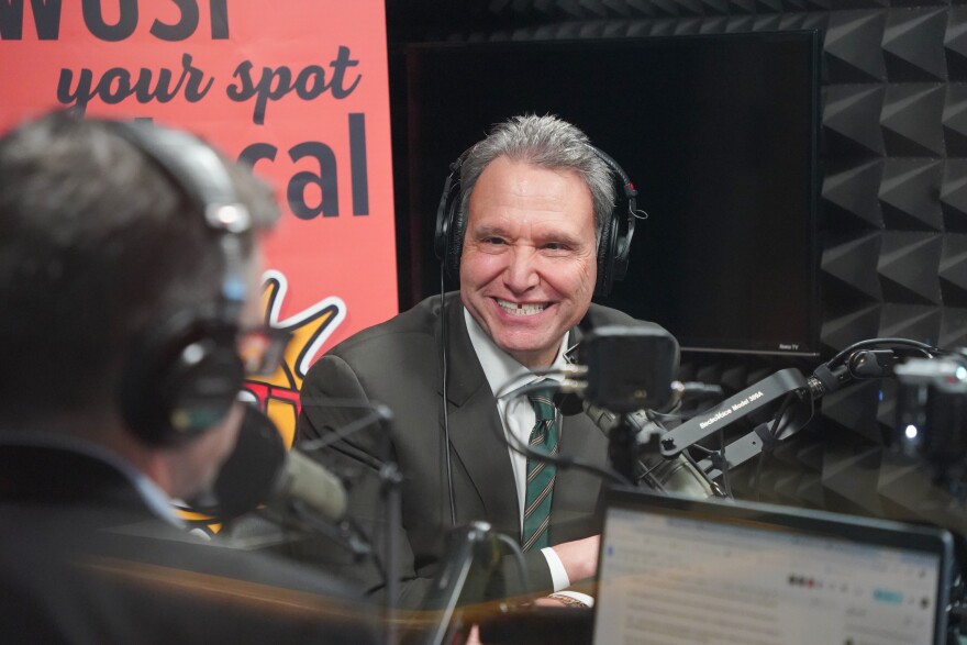 Man in dark green suit and tie smiles as he sits by a microphone in a studio 