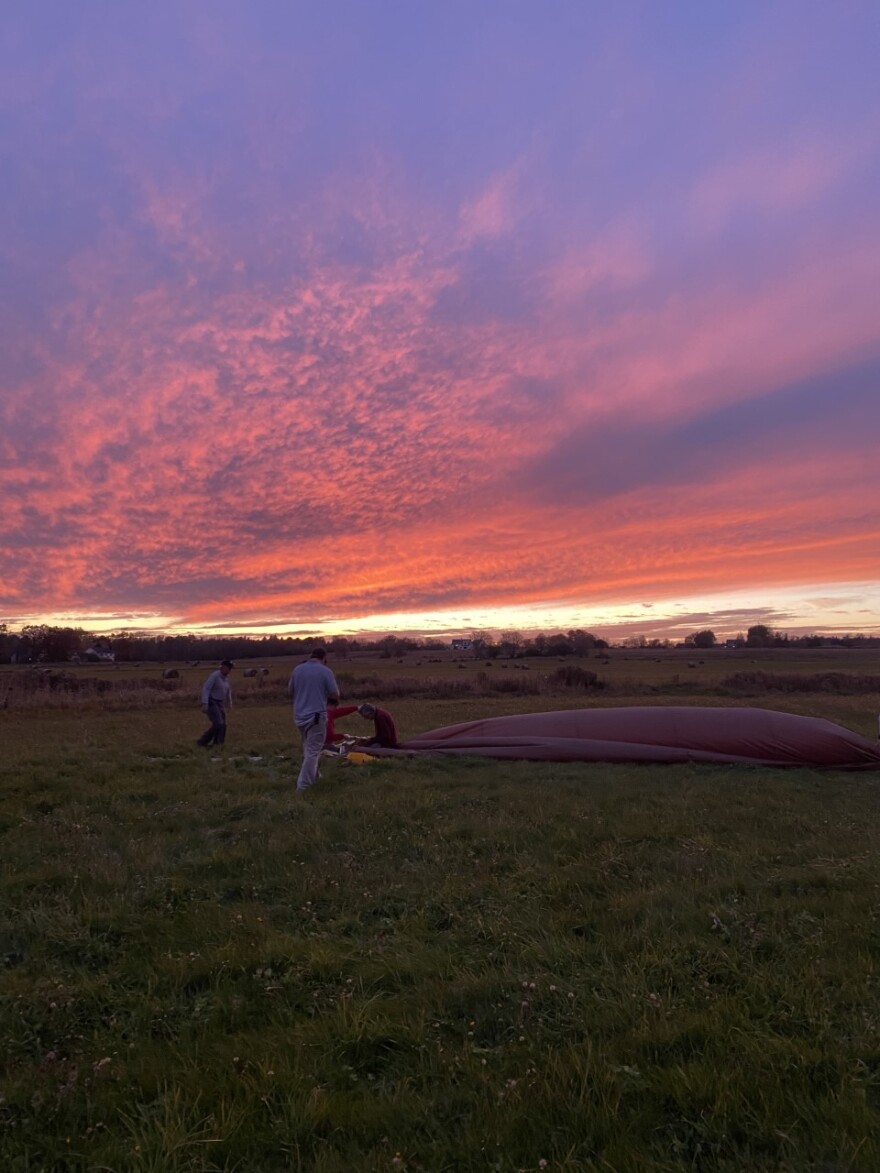 A photo of a deflated hot air balloon in a field at sunset when the sky is bright pink and deep purple. People are milling about the field. 