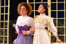 Two women in Victorian dresses stand arm in arm, scoffing at one another