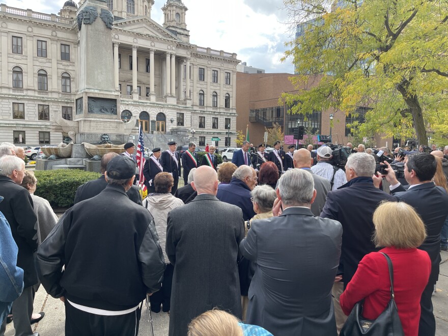 A crowd of multiple people stands together and surrounds the Columbus Monument which stands in the center of a fountain.