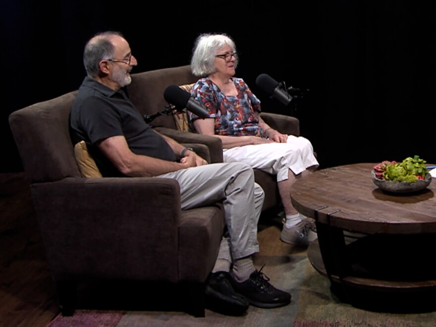 A smile couple sits on arm chairs in front of microphones, Ed Deller on the left and Joanne Newcomb-Losh on the right