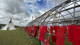 Red dresses hanging on a wooden structure and on the left in the background, there is a teepee. 