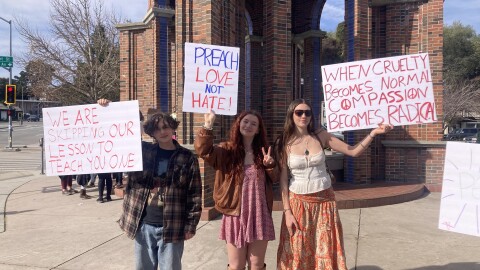 Three high school students hold signs in front of the Santa Cruz Clocktower. From left to right they read: "We are skipping our lesson to teach you one," "Preach love not hate!" and "When cruelty becomes normal compassion becomes radical".