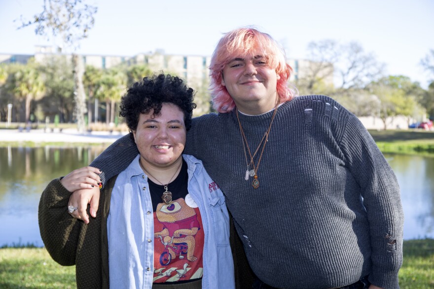 A young afro latino trans man  with short, curly dark hair and wearing a blue button up shirt stands next to a white trans woman with pink hair and a great sweater. They're both smiling.