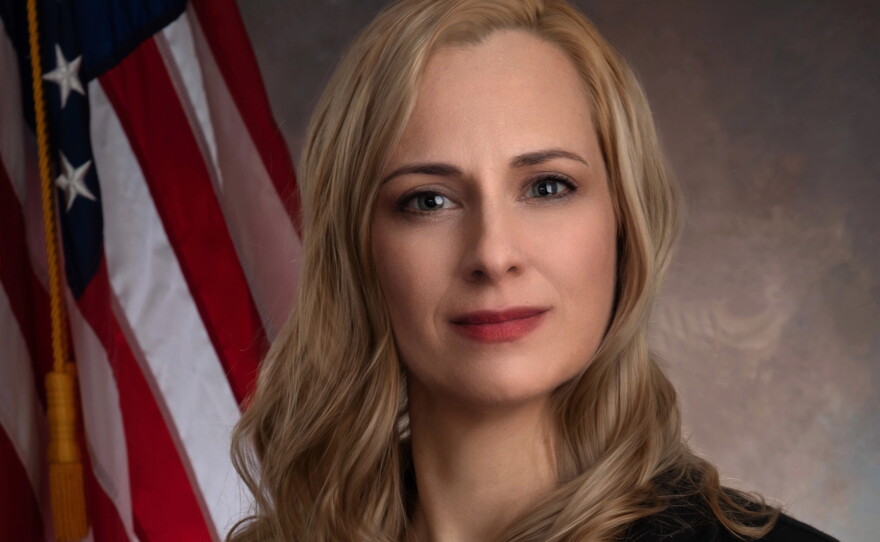 A woman with long blonde hair is wearing a black blazer over a red blouse and posing next to an American flag.