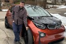 Tyler Lake in a brown chore shirt and jeans standing next to a reddish-brown and black BMW car in a concrete driveway. One hand is in his pocket, one is grabbing a front tire. View of a suburban neighborhood with lingering snow is visible in the background. 