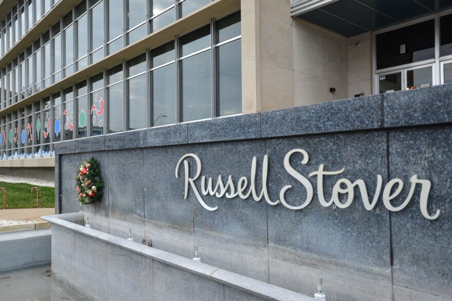 A granite wall with a Christmas wreath sits outside the front of a building. On the granite wall is a sign in script letters spelling "Russell Stover." Behind the wall is a large building with holiday decorations on the front.