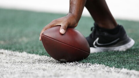 A close up shot of an unidentifiable person off screen grabbing a football. The brown oval shaped ball is in the center of the image, pressed against the ground by a hand whose arm cuts off right above the wrist. The person's foot can also be seen in the image, just behind the ball. Their wearing a black Nike tennis shoe with the white outline of Nike's check mark symbol imprinted on the side of the shoe. 