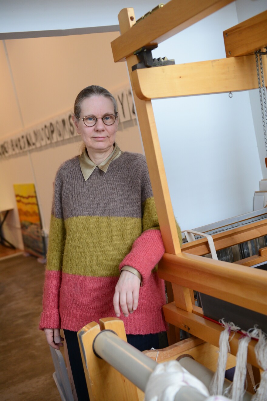 A woman in a colorful sweater stands by a large loom