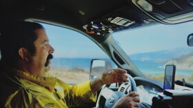A man in a yellow firefighting jacket drives on a highway with the ocean behind him. 